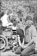 photo shows the long-haired Wade Blank seated on the ground to talk to a woman who's using a wheelchair.