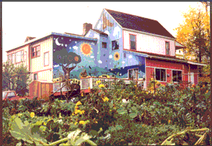 the postcard shows a riot of a colorful house that's painted with a mural and has a couple of red porches and is surrounded by a riot of a garden full of blooming sunflowers.